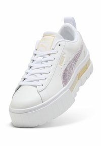 Balts Puma sporta apavu pāris ar biezu zoli, gaiši violetu zamša svītru, bēšiem Puma logotipiem un baltām šņorēm, no leņķa, kas rāda priekšpusi un sānu.