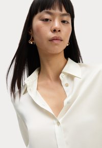 Blusa blanca de seda con un cuello estructurado y cierre frontal de botones. La modelo lleva pendientes dorados y llamativos. Textura suave y ajuste relajado.