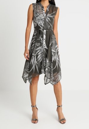 Robe sans manches noire et blanche à fleurs avec patte de boutonnage devant, taille cintrée par une ceinture, et ourlet en forme de mouchoir, portée avec des talons à bride métallique à la cheville.