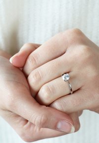 Zilveren ring met een enkele ronde geslepen diamant, gladde band en klauwzetting. Handen in elkaar verstrengeld, waarmee de prominentie van de ring wordt benadrukt.