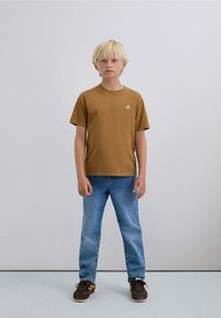 Camiseta marrón de manga corta con un pequeño logo, combinada con jeans celestes y zapatillas oscuras, en un niño de pie con cabello rubio claro.