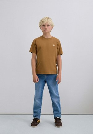 Camiseta marrón de manga corta con un pequeño logo, combinada con jeans celestes y zapatillas oscuras, en un niño de pie con cabello rubio claro.