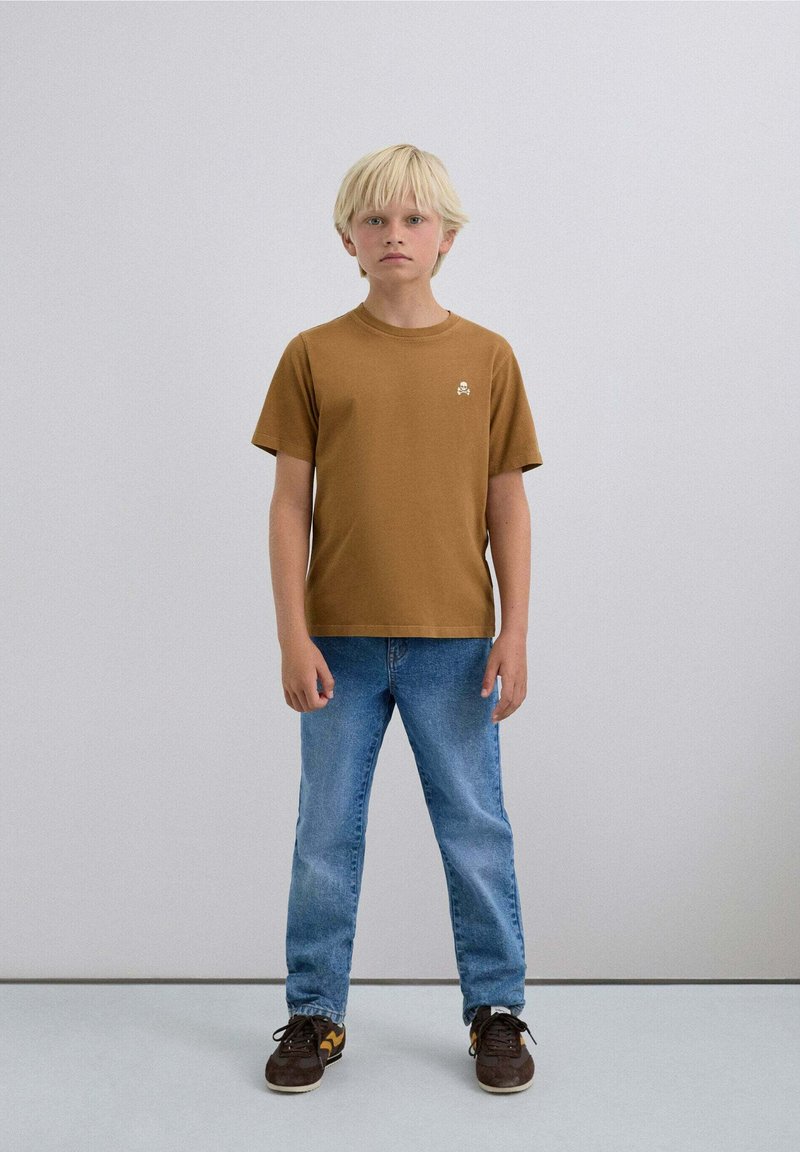 Camiseta marrón de manga corta con un pequeño logo, combinada con jeans celestes y zapatillas oscuras, en un niño de pie con cabello rubio claro.