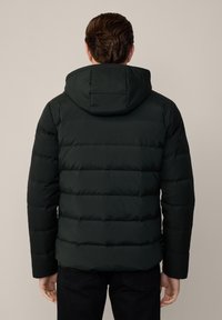 Uomo che indossa una giacca a cappuccio nera imbottita e pantaloni neri, visto da dietro su uno sfondo chiaro e semplice.