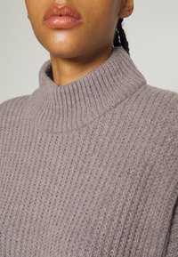 Grå stickad tröja med hög ribbad krage, som har ett texturerat, mjukt tyg och en avslappnad passform. Närbild av halsringningen och övre bröstområdet.