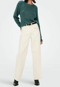 Pull en tricot vert avec texture côtelée, associé à un jean crème à taille haute et jambes larges, et une ceinture noire. Des bottines noires complètent la tenue.