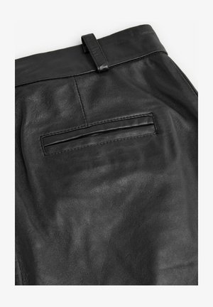 Mustad nahkpüksid, millel on sile tekstuur, taga tasku klapiga ja vöölink pahkluul.