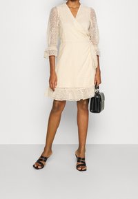 Robe enveloppante de couleur crème avec des manches longues transparents, tissu texturé, ourlet à volants, associée à un petit sac à main noir et des sandales à glisser noires.