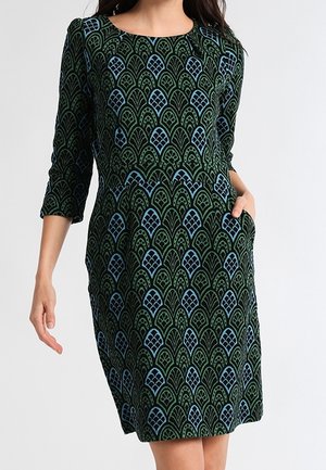 Robe longueur genoux avec manches 3/4 présentant un motif écailles vert et bleu, portée avec une main dans la poche sur un fond uni.
