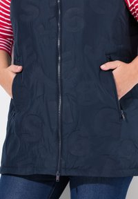 Námořnická bezrukávová puffer vesta se zipem vpředu, bočními kapsami a strukturovaným vzorem písmen a čísel. Hladký, lehký materiál.