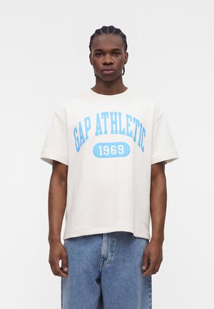 Νέος άνδρας με κοτσίδες που φοράει λευκό φαρδύ T-shirt με μπλε γράμματα "GAP ATHLETIC 1969" και άνετο μπλε τζιν.