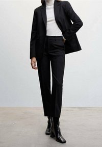 Mustavalmistettu puku, jossa on sopiva bleiseri ja suorat housut, yhdistettynä vaaleaan neuleeseen ja kiiltävät mustat saappaat, joissa on teksturoitu kuvio.