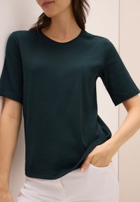 Tmavě zelená košile s krátkým rukávem a jemně strukturovaným povrchem, kulatým výstřihem a volným střihem, doplněná světlými kalhotami.