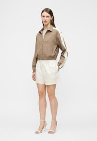 Γυναίκα με καφέ μπουφάν με φερμουάρ και λευκές ρίγες στα μανίκια, κρεμ σορτς με καρό σχέδιο και μπεζ πέδιλα με ψηλό τακούνι, στέκεται μπροστά σε λευκό φόντο.