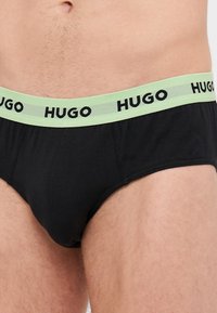 Culottes en coton noir avec une ceinture élastique vert clair arborant "HUGO" en lettres noires. Texture lisse, design taille basse.