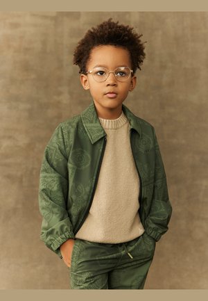 Veste à motifs verts portée sur un pull beige, associée à un pantalon vert assorti. Les accessoires comprennent des lunettes rondes et des poignets élastiques.