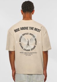 Beige oversized t-shirt med et rygdesign med fed sort tekst og et grafik af en due med udfoldede vinger, lavet af blødt bomuldsstof.