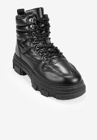 Botas de combate negras con parte superior acolchada, acabado de cuero brillante, cordones en la parte delantera y suela de goma gruesa con surcos profundos para tracción.