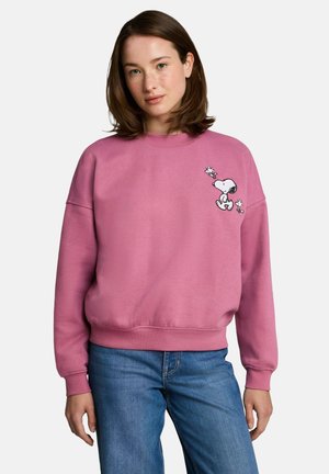 Junge Frau mit braunen Haaren trägt einen pinken Pullover mit Snoopy- und Woodstock-Motiven, kombiniert mit blauen Jeans.