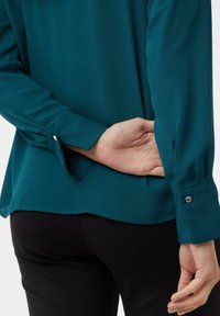 Teal blouse with long sleeves, curved hem, and buttoned cuffs. Smooth fabric with a sleek appearance, paired with black pants.