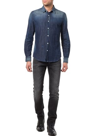 Camisa vaquera azul oscuro con cuello abotonado, mangas largas y un sutil desgaste. Combinada con vaqueros gris oscuro y zapatos negros.