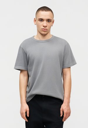 Νεαρός με κοντά μαλλιά, που φοράει μια απλή γκρι κοντομάνικη μπλούζα και μαύρο παντελόνι, στέκεται μπροστά σε λευκό φόντο.