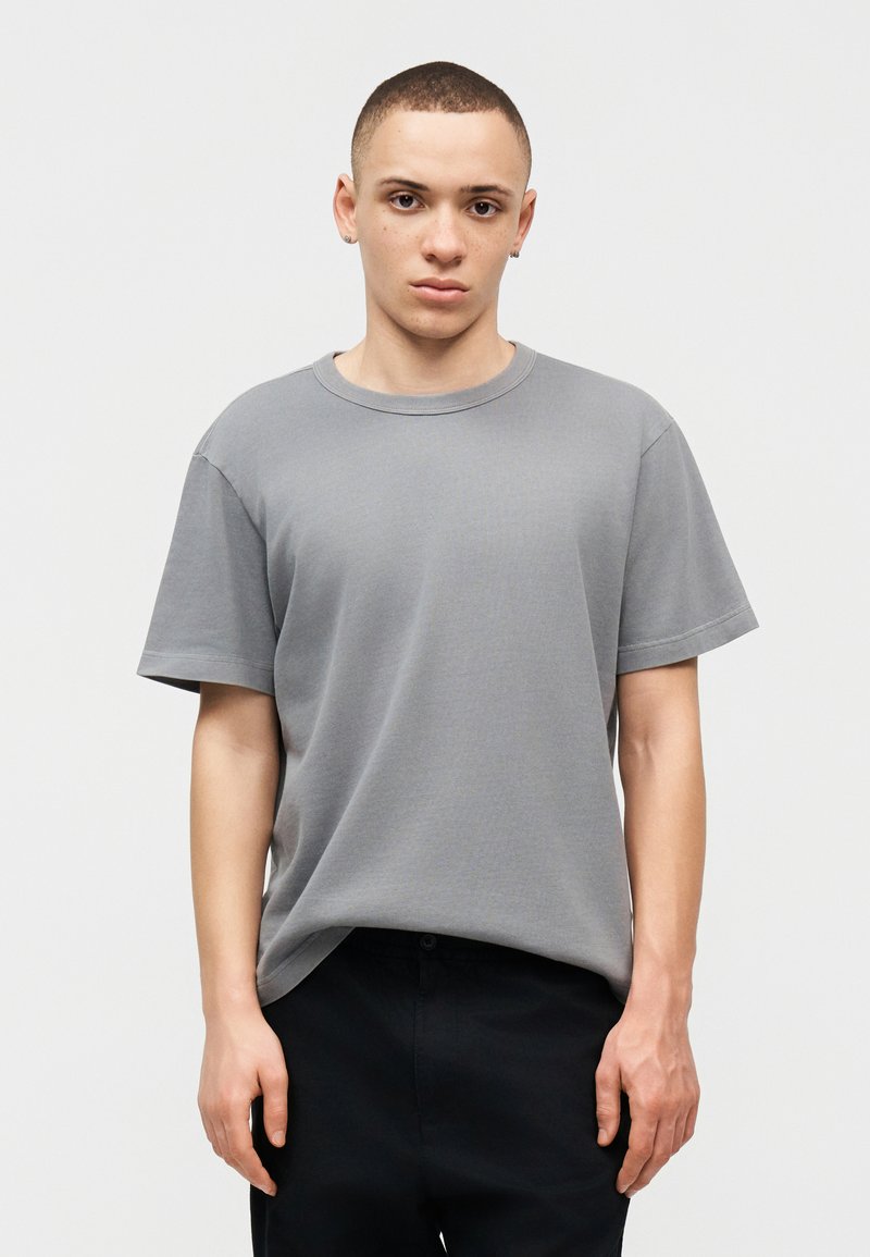 Jeune personne aux cheveux courts portant un t-shirt gris uni à manches courtes et un pantalon noir, debout devant un fond blanc.