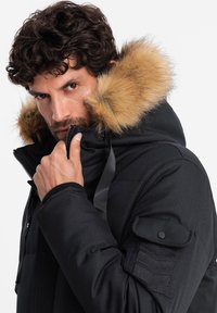 Parka invernale nera con cappuccio in pelliccia sintetica, chiusura con zip e polsini a coste. Presenta toppe sulle spalle e una vestibilità strutturata.