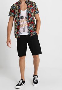 Camicia a maniche corte floreale con motivi verdi e rossi su sfondo nero, canottiera bianca con grafiche, pantaloni corti neri e sneakers nere.