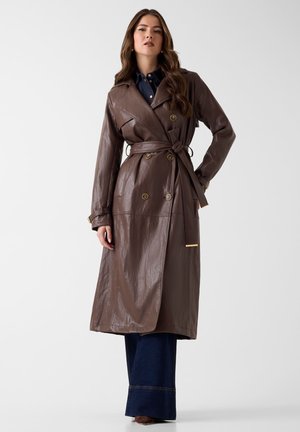 Kvinde iført en brun læder trenchcoat med bælte over mørke denimbukser, stående med den ene hånd på hoften foran en ensfarvet baggrund.