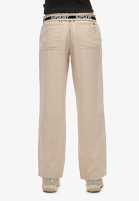 Pantaloni beige in lino a gamba larga con una vestibilità rilassata, dotati di due tasche posteriori e una cintura elastica con marchio in blu e bianco.