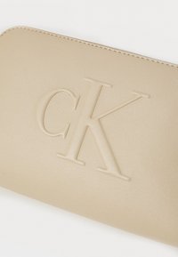 Bež novčanik od umjetne kože s utisnutim "CK" logom, glatke teksture, zaobljenih rubova i kontrastnim šavovima. Kompaktni dizajn za laku upotrebu.