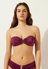 Conjunto de bikini en un profundo morado, que incluye un top tipo bandeau con tela fruncida y un detalle de hardware en forma de U dorado, acompañado de bragas fruncidas.