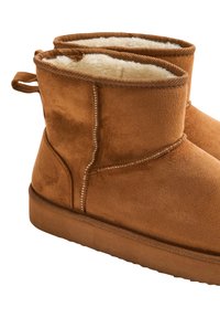 Stivaletti alla caviglia in camoscio marrone con fodera morbida color crema, suola piatta e cucitura verticale lungo il lato. Anello sul retro per facilitarne la calzata.