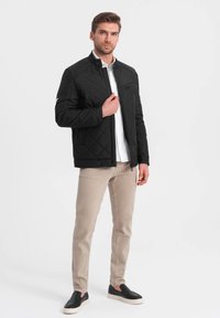 Sort quiltet jakke med opretstående krave, frontlynlås og sidelommer, båret over en hvid skjorte, parret med lysebeige bukser og sorte sko.