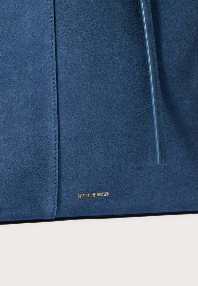 Borsa a tracolla in suede blu con texture liscia, dotata di una tracolla trasparente a contrasto e un logo dorato stampato alla base della borsa.