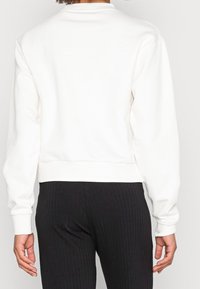 Valkoinen puuvillainen collegepaita, jossa resorireunus helmassa ja hihansuissa; pyöreä pääntie; yksinkertainen, kuvioimaton malli; malli pukeutunut mustiin resoreihin.