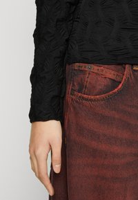 Zwarte textuur top met een gerimpeld patroon, gecombineerd met rode spijkerbroek met gele stiksels en een detail aan de voorkant van de zak.