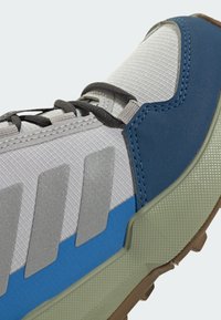 Sportovní obuv se šedým síťovaným svrškem, modrými syntetickými překryvy, reflexními stříbrnými prvky a texturovanou zelenou gumovou podešví.