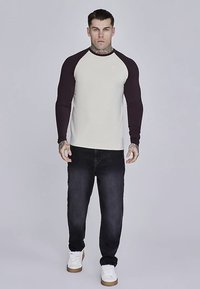 Maglia a maniche lunghe color crema con maniche bordeaux scuro, abbinata a jeans scuri e sneaker bianche. Presenta uno scollo rotondo e un design aderente.