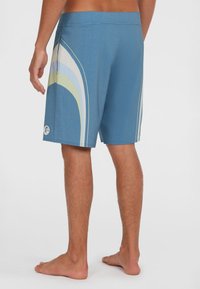 Blaue Badeshorts aus dehnbarem Material, mit einem Retro-Wellenmuster in Weiß, Hellblau und Grün, und einer Seitentasche.