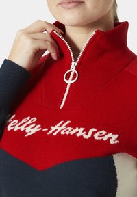 Suéter de punto rojo y azul marino con un alto cuello y cremallera, que presenta un destacado logo blanco en el pecho y bloques de color contrastantes.