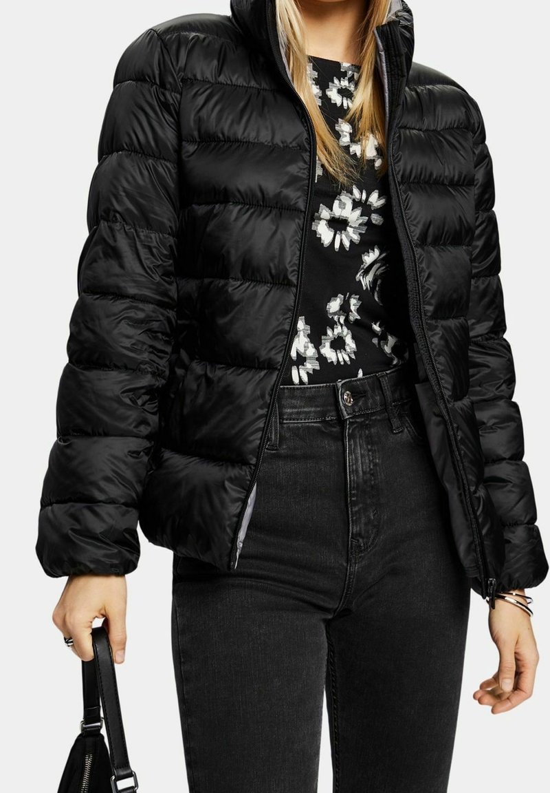 Femme portant une doudoune noire par-dessus un haut noir à fleurs, rentrée dans un jean noir taille haute, tenant un sac à main noir.