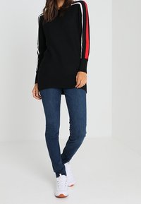 Kvinna i en svart långärmad tröja med röda och vita ränder; bär mörka jeans och vita sneakers, stående mot en ljus bakgrund.