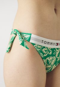Grønn bikiniundertøy med blomstermønster, som har en sløyfe på siden og en kontrasterende hvit midjebånd med merke.