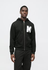 Μαύρο φούτερ με φερμουάρ από βαμβακερό μείγμα, διαθέτοντας μπροστινή τσέπη, κουκούλα με κορδόνι και γραφικό λογότυπο σε λευκή ετικέτα.