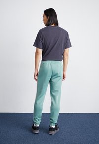 Herrenoutfit bestehend aus einem dunkelgrauen T-Shirt mit kurzen Ärmeln und hellgrünen Jogginghosen mit weißen Seitenstreifen. Die Hose hat einen elastischen Bund.