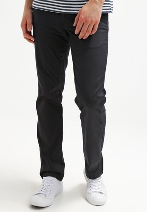 Homme portant un pantalon noir slim, des baskets blanches et une chemise rayée bleu marine et blanche, debout devant un fond blanc.