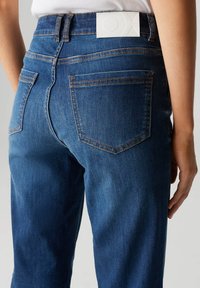 Jeans in denim blu scuro con silhouette aderente, tasche posteriori classiche e un'etichetta in pelle bianca con logo impresso sulla cintura.