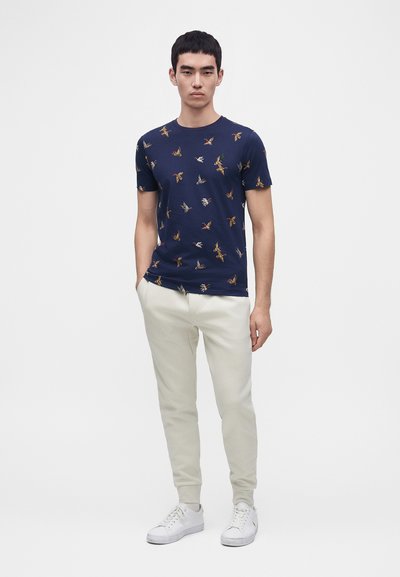 Νέος άνδρας που φοράει μπλε ναυτικό t-shirt με σχέδια πουλιών, ανοιχτόχρωμο παντελόνι jogger και λευκά αθλητικά παπούτσια, στέκεται με το ένα χέρι στην τσέπη.